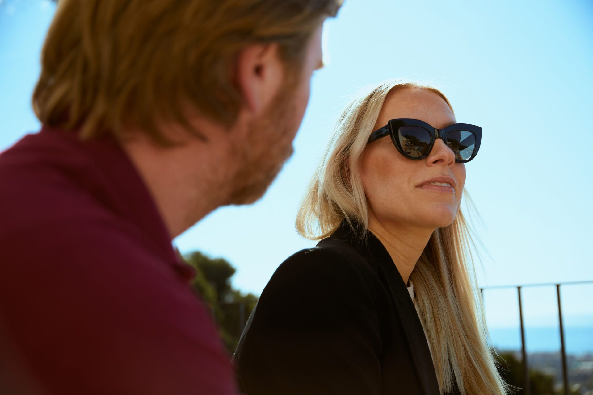 Två personer som samtalar. I förgrunden en person med vinröd pike och i fokus en person i svart kavaj och svarta solglasögon.