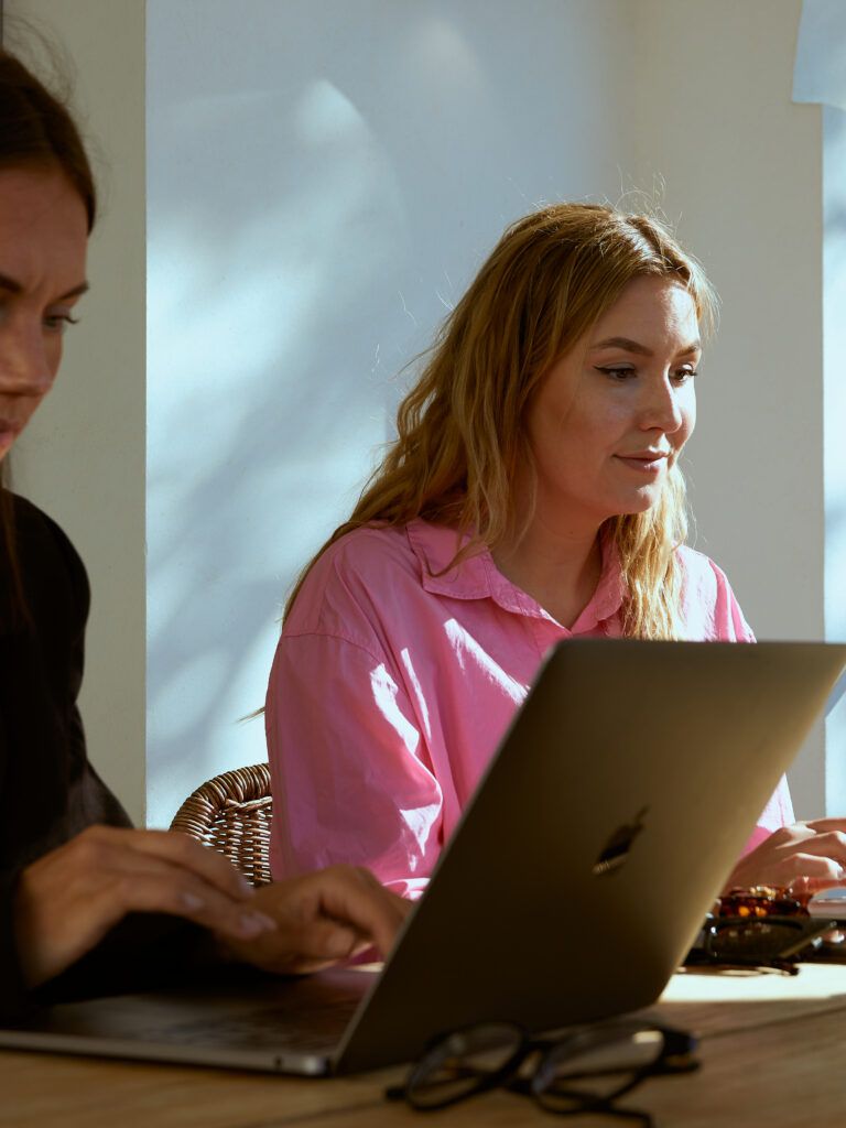 Martina Myrén och Alexandra Åhre sitter bredvid varandra med varsin laptop framför sig som dom arbetar på. De sitter vid ett konferensbord på en veranda och i bakgrunden ser man en blå himmel och gröna träd. Alexandra har på sig en rosa skjorta, medan Martina har en svart kavaj med vit tröja därunder.