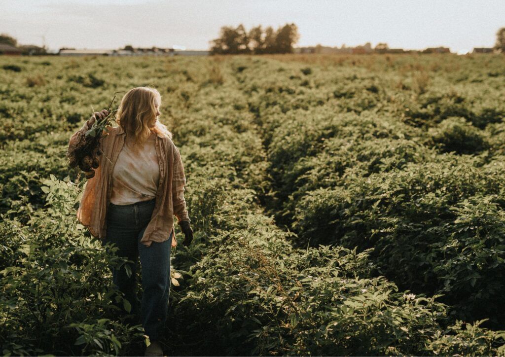 En blond kvinna står utomhus mitt i ett potatisodlingsfält under sommartid.