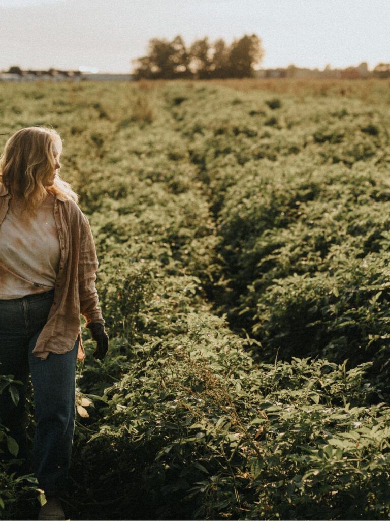 En blond kvinna står utomhus mitt i ett potatisodlingsfält under sommartid.