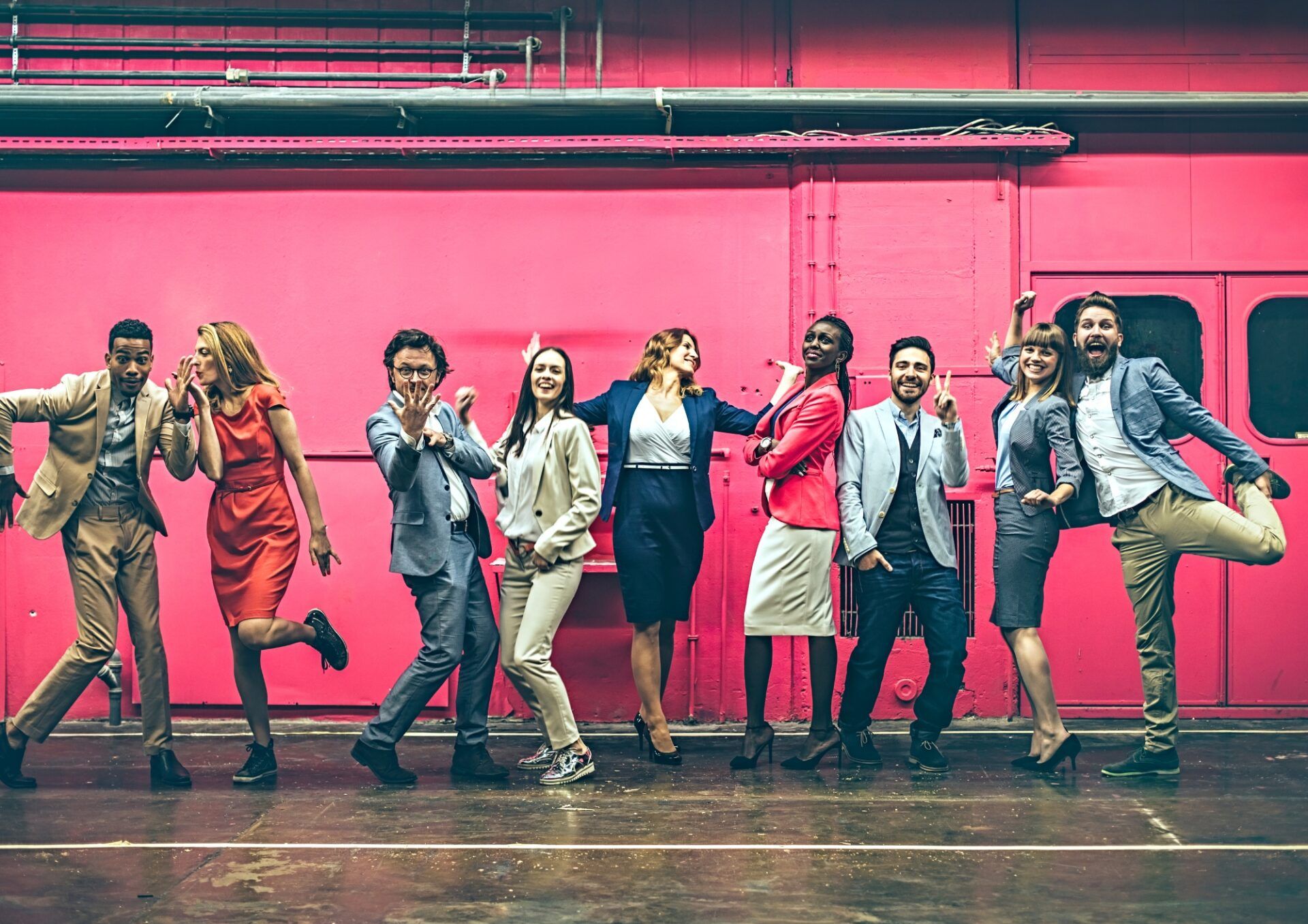 En grupp med människor i kontorskläder som står framför en rosa industrivägg. De dansar och ler.