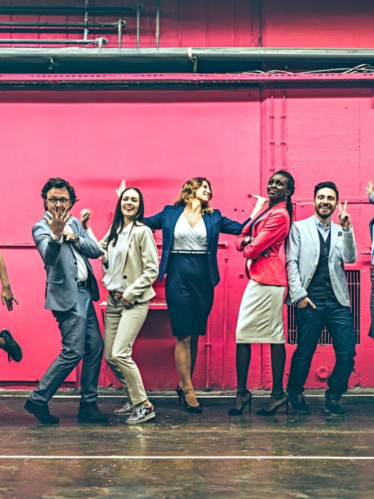 En grupp med människor i kontorskläder som står framför en rosa industrivägg. De dansar och ler.