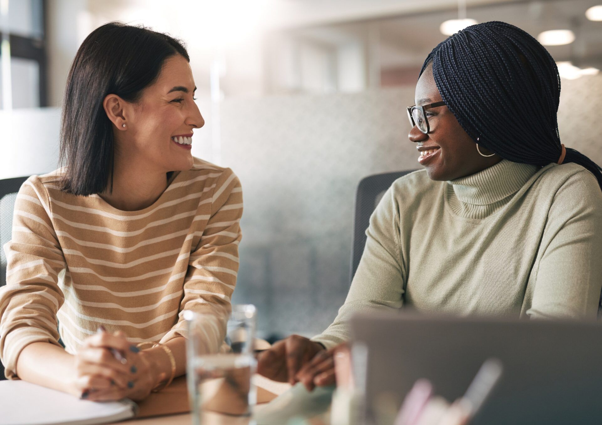 Två leende kvinnor sitter vid ett skrivbord i en kontorsmiljö och har en konversation. De är vända mot varandra