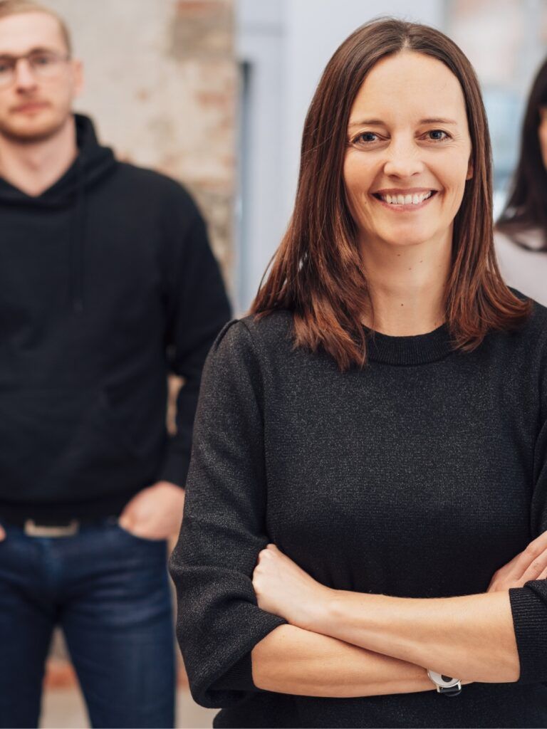 I förgrunden syns en kvinna som ler och har brunt, axellångt hår. I bakgrunden står hennes team, bestående av fyra personer. Alla är ledigt klädda och ser in i kameran.