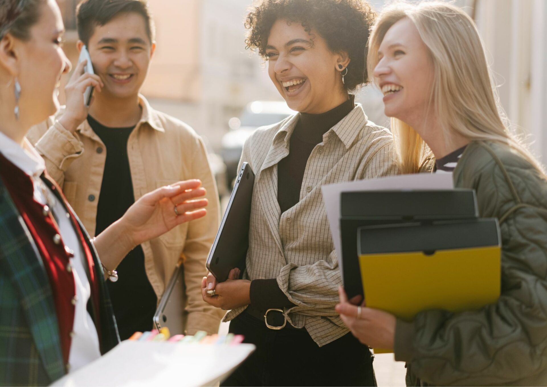 Fyra personer står utomhus och skrattar med varandra. De bär på datorer och pärmar.