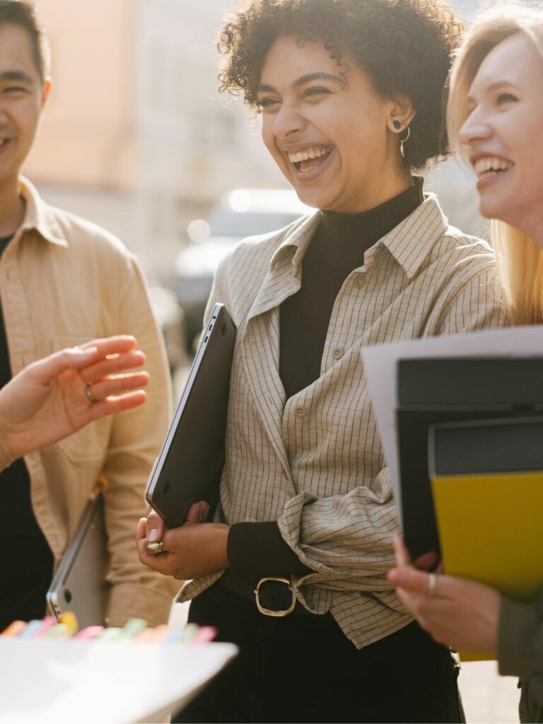 Fyra personer står utomhus och skrattar med varandra. De bär på datorer och pärmar.