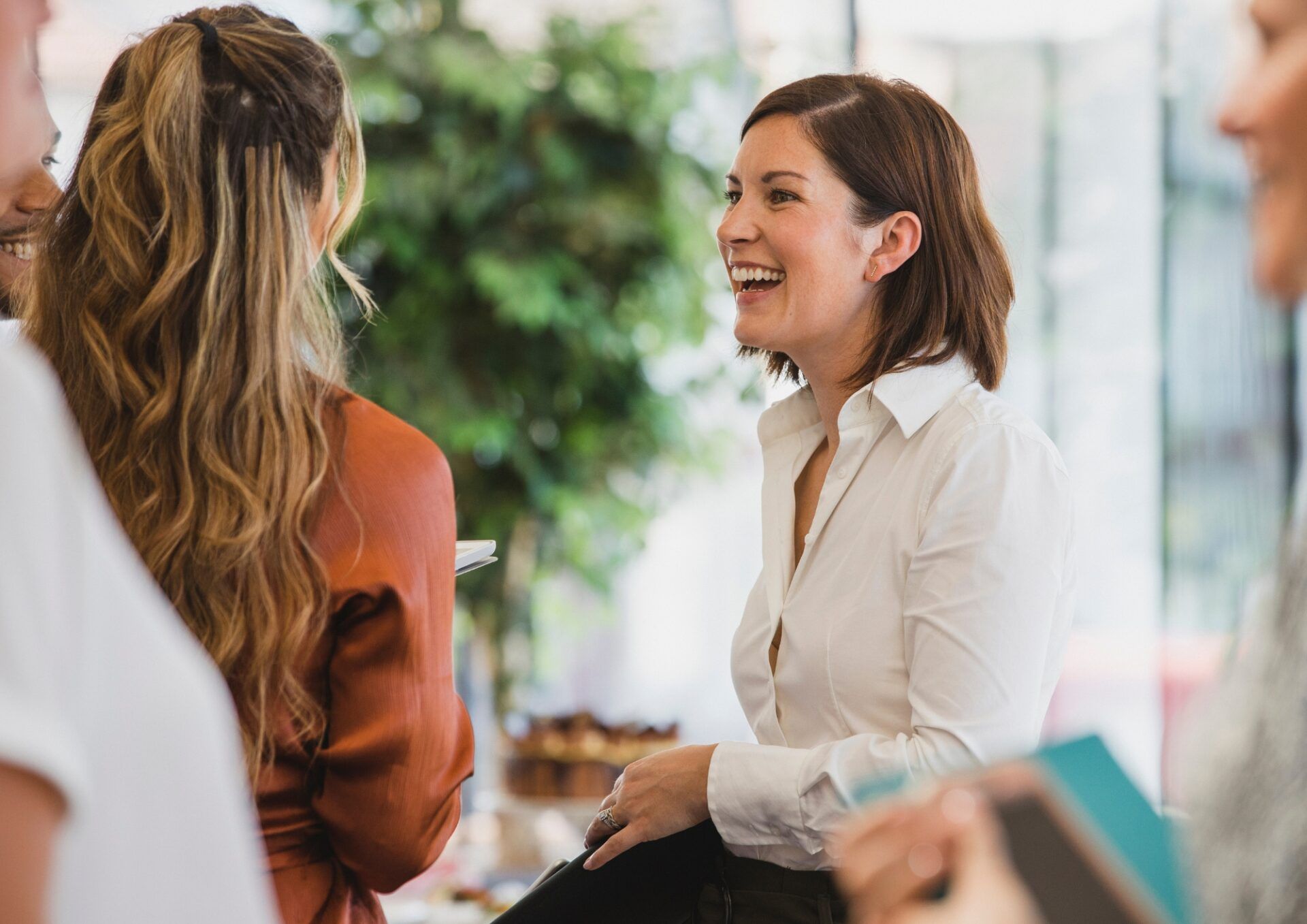 bild på en person med mörkt axellångt hår i en vit skjorta. Personen står vänd mot en annan person och skrattar.