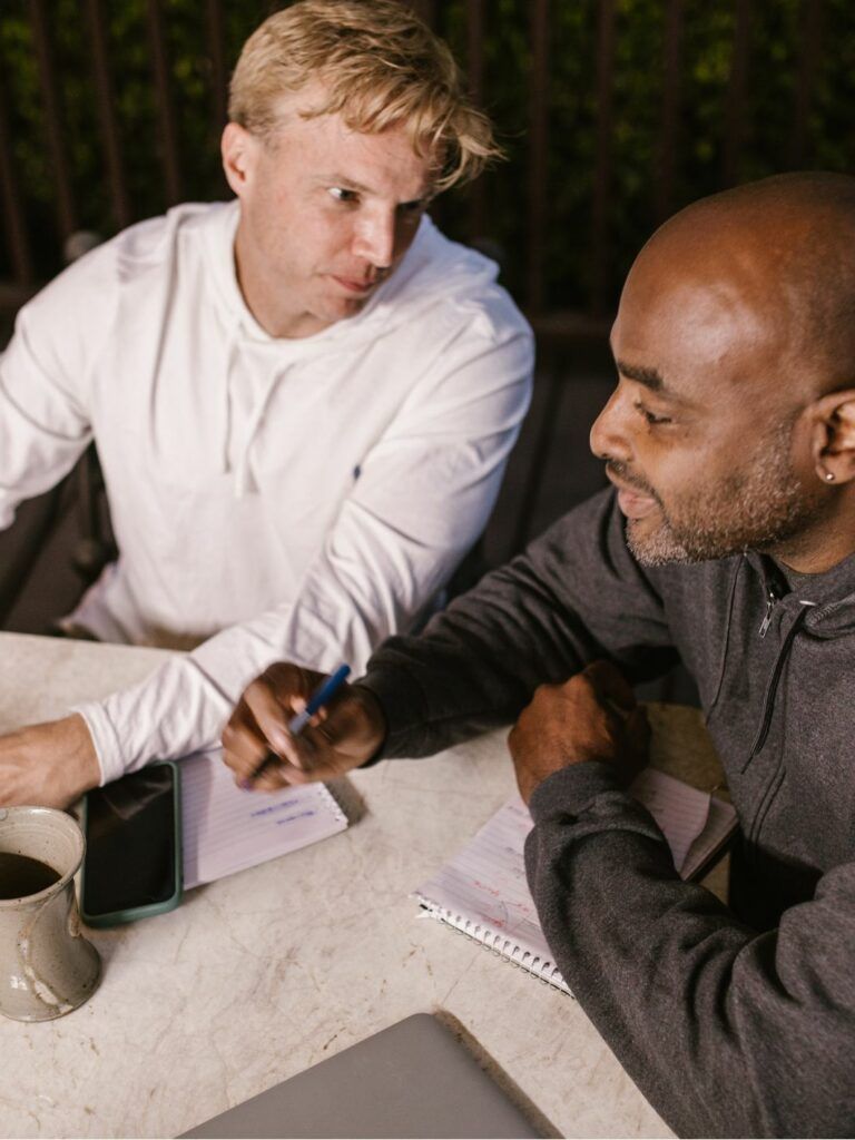 Två män sitter bredvid varandra vid ett skrivbord, framför en dator och samtalar.