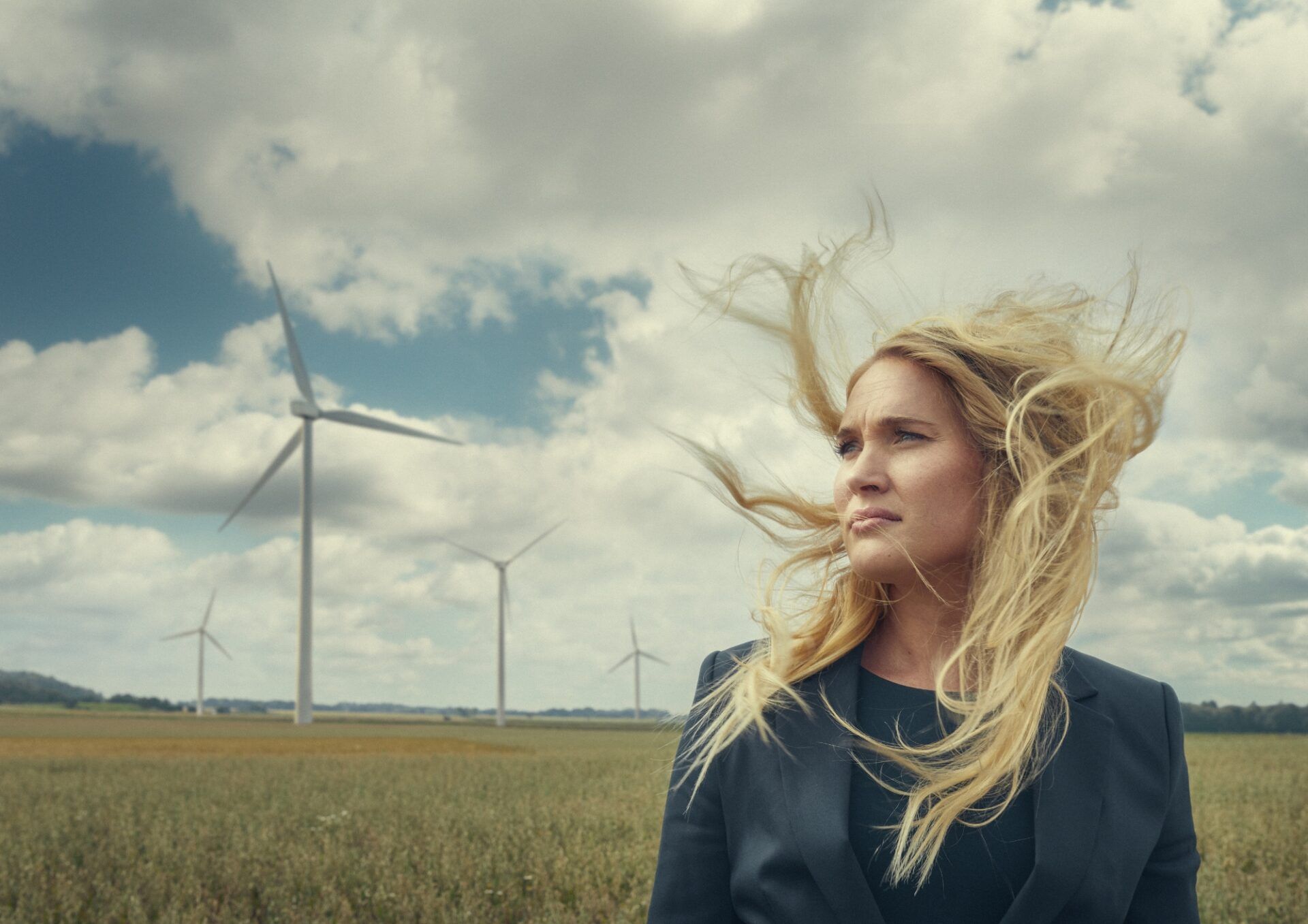 En ung jurist står på ett sädesfält med håret blåsande i vinden. Hon blickar ut över landskapet. I bakgrunden syns ett antal vindkraftverk.