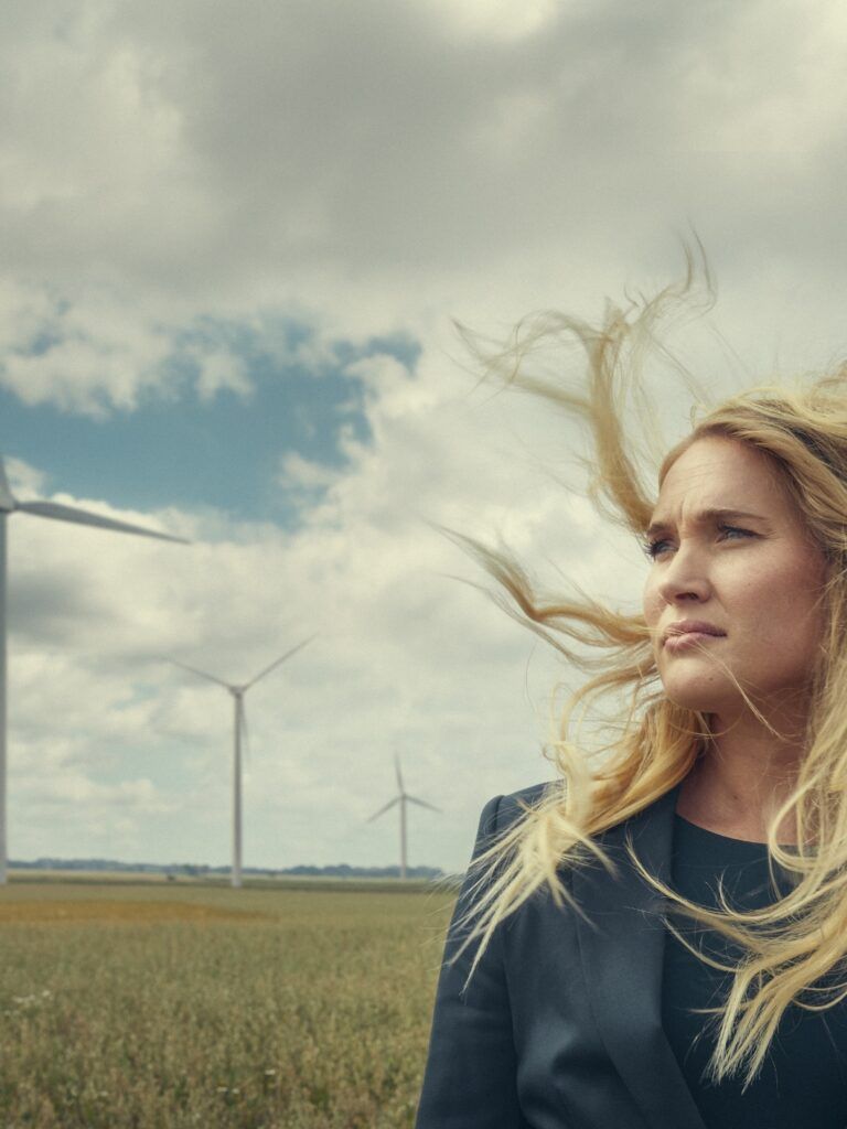 En ung jurist står på ett sädesfält med håret blåsande i vinden. Hon blickar ut över landskapet. I bakgrunden syns ett antal vindkraftverk.