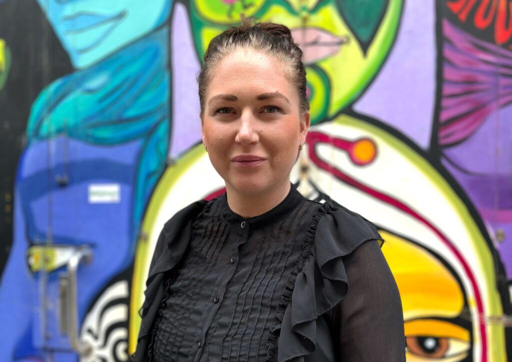 Foto på Carolina Elofsson. Carolina har håret upsatt i en knut och bär svart blus. I bakgrunden syns en färgglad grafittivägg.