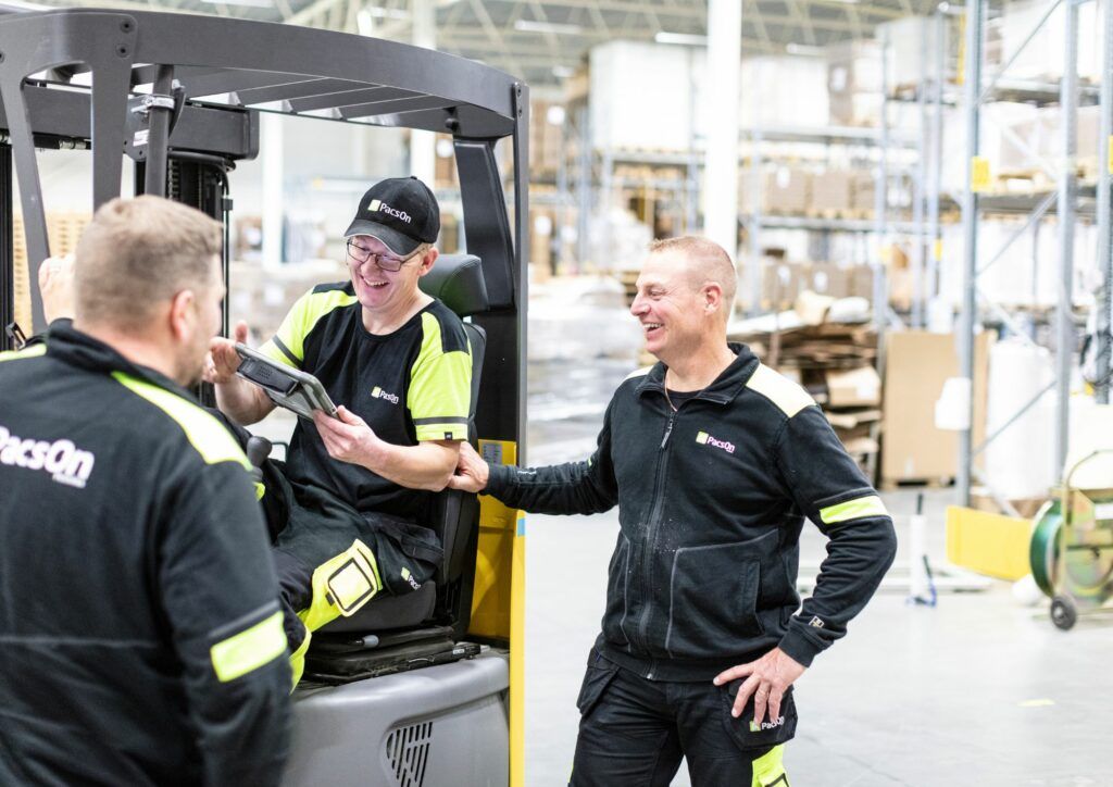 Tre personer som bär arbetskläder i lagermiljö. Två står bredvid en truck och den tredje sitter i trucken och visar något på en platta.