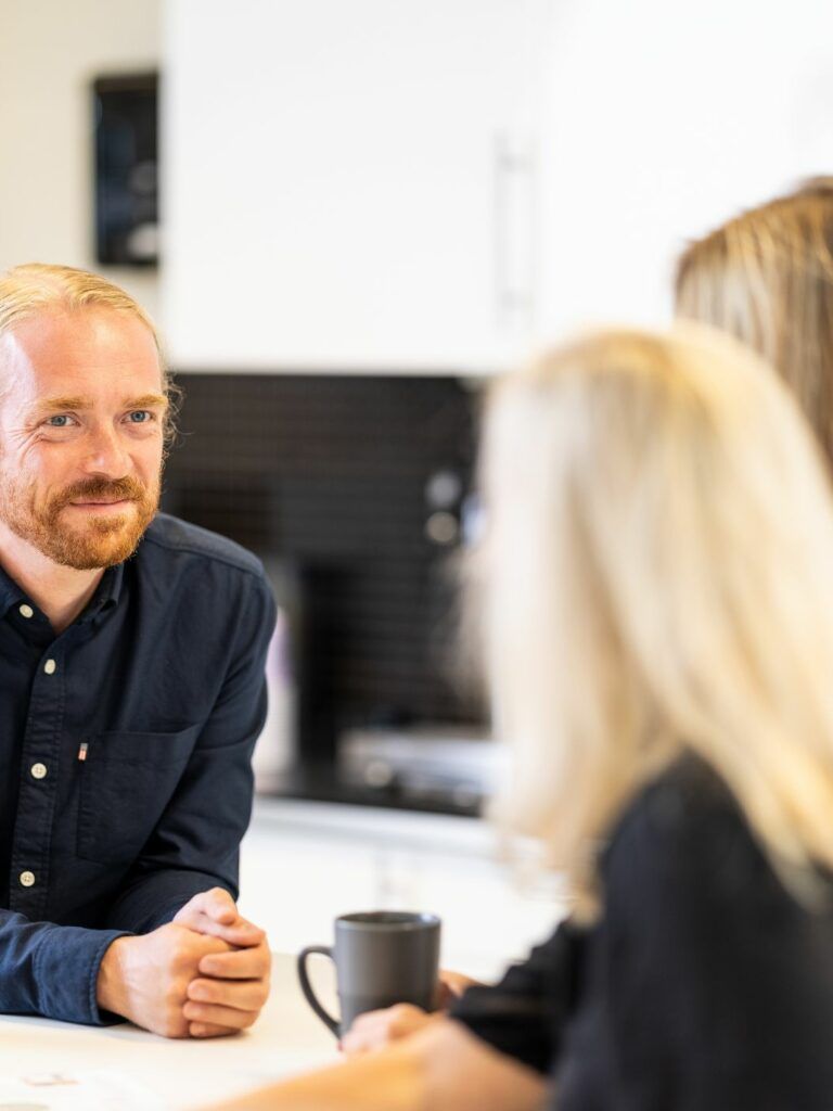 I förgrunden ryggen av två personer med halvlångt blont hår. I fokus en person med mörkblå skjorta, blåa ögon och tillbakakammat blont hår.