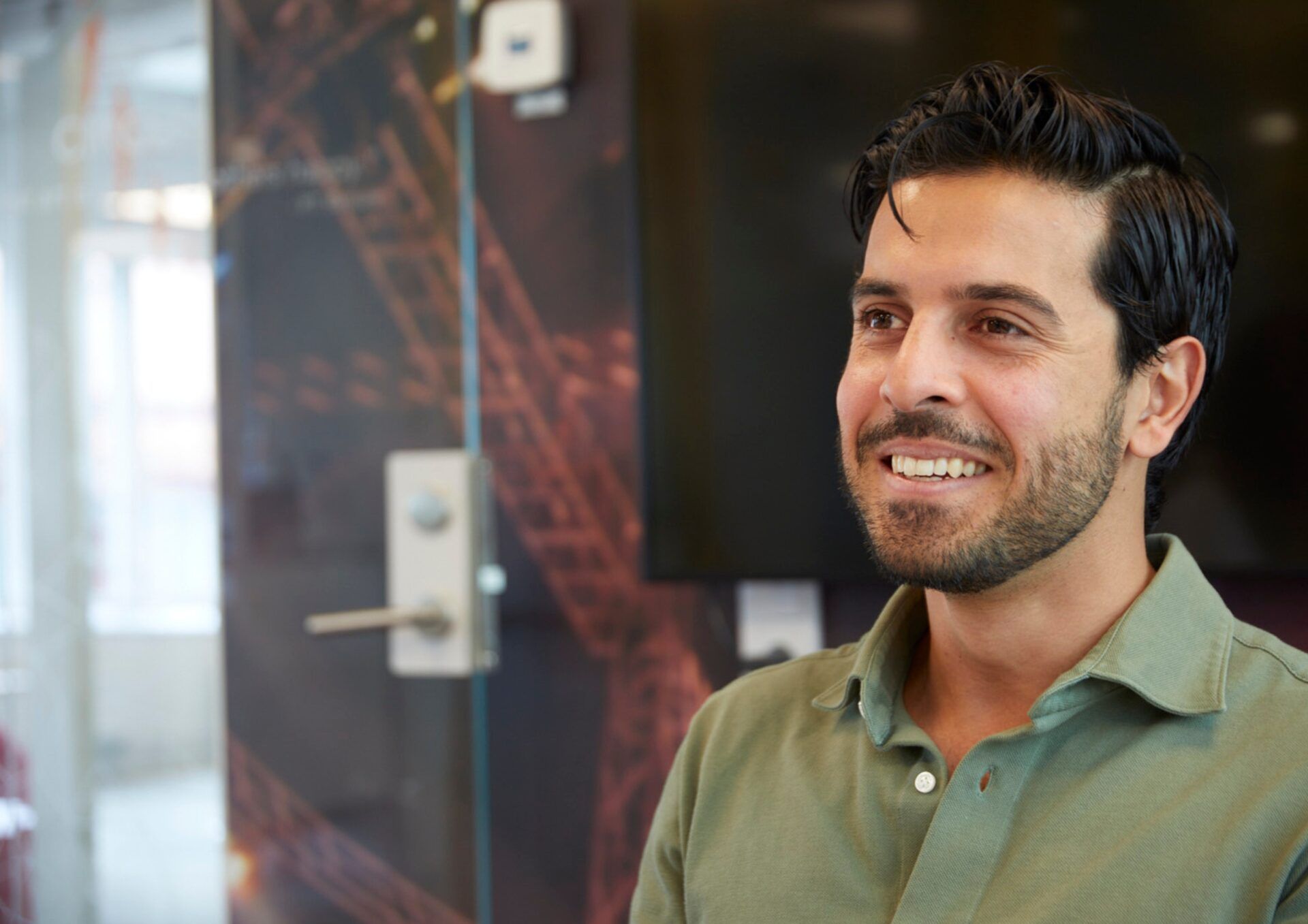 Poyan Karimi ler stort i en olivgrön skjorta. Han befinner sig i en ljus kontorsmiljö.