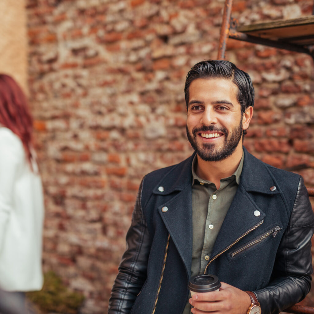 Poyan Karimi ler stort framför en tegelvägg. Bakgrunden är suddig, man ser endast lite av en rödhårig tjej. Poyan har på sig en grön skjorta med skinnjacka över, samt håller en kaffemugg i handen.