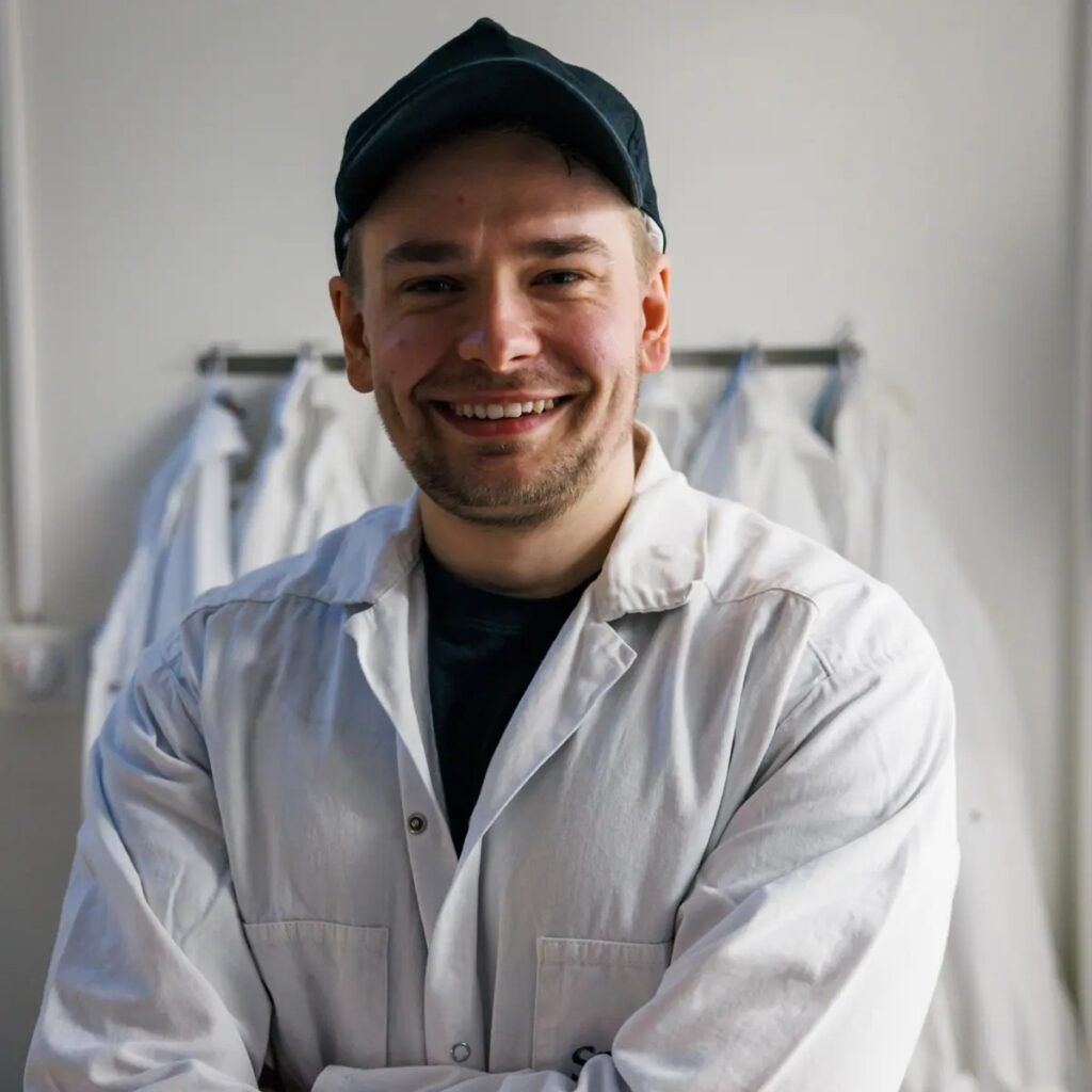 Person med labbrock och keps som tittar in i kameran och ler