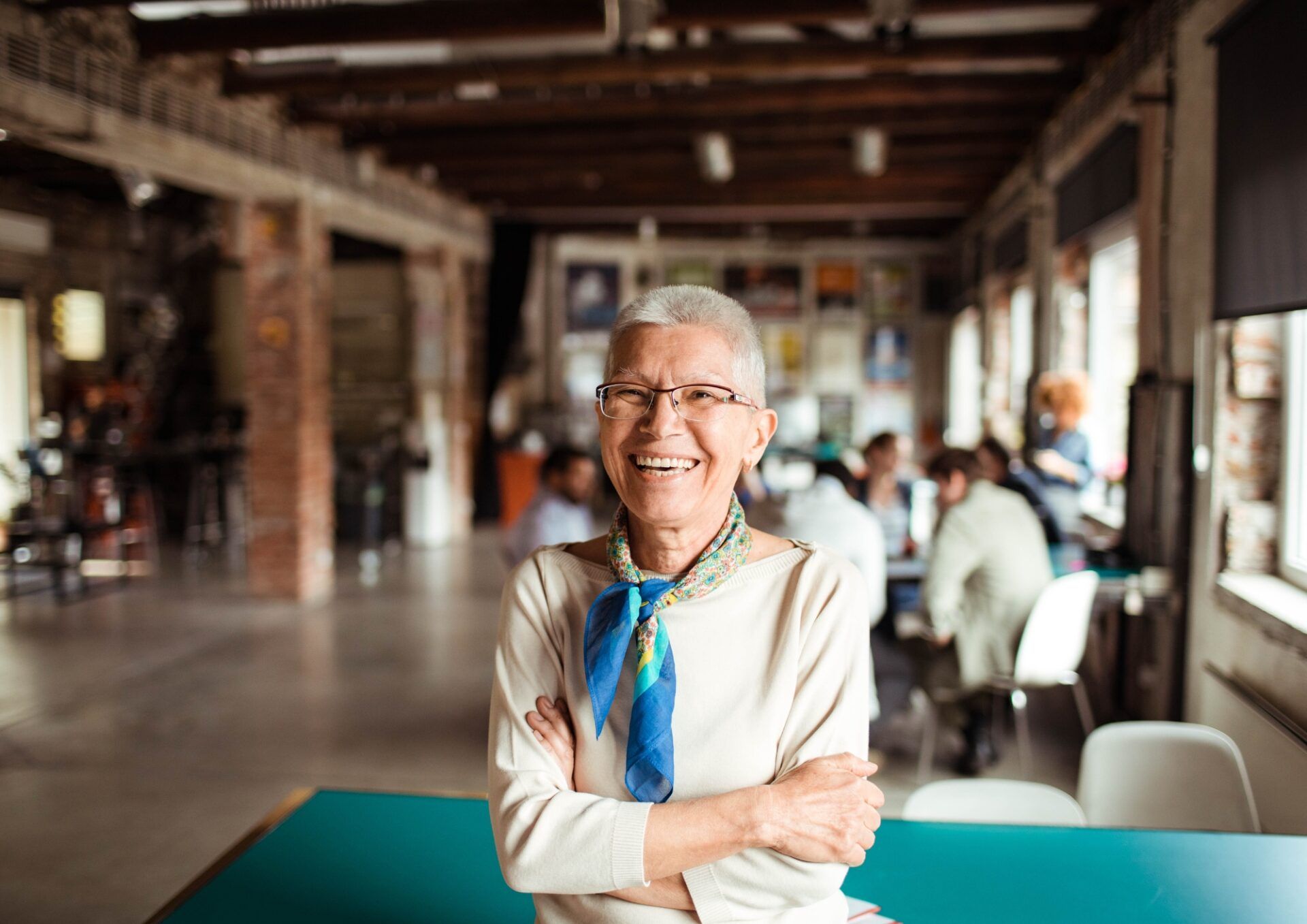 Leende äldre kvinna med glasögon och blå scarf står med armarna i kors i en ljus, modern lokal.