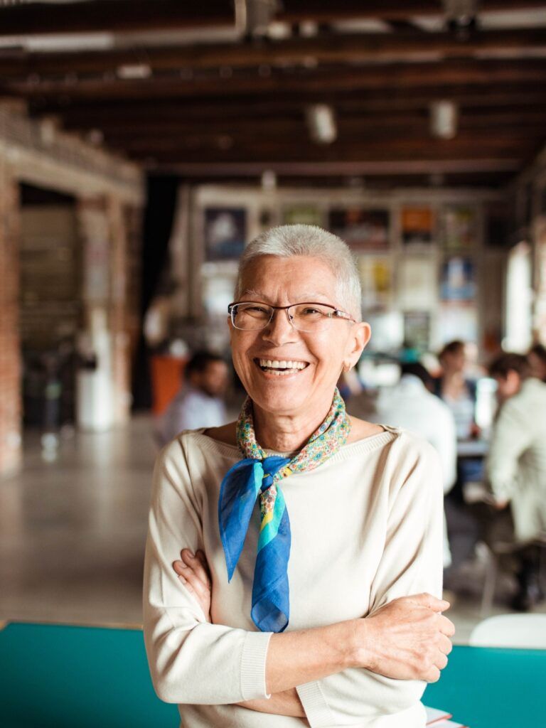 Leende äldre kvinna med glasögon och blå scarf står med armarna i kors i en ljus, modern lokal.