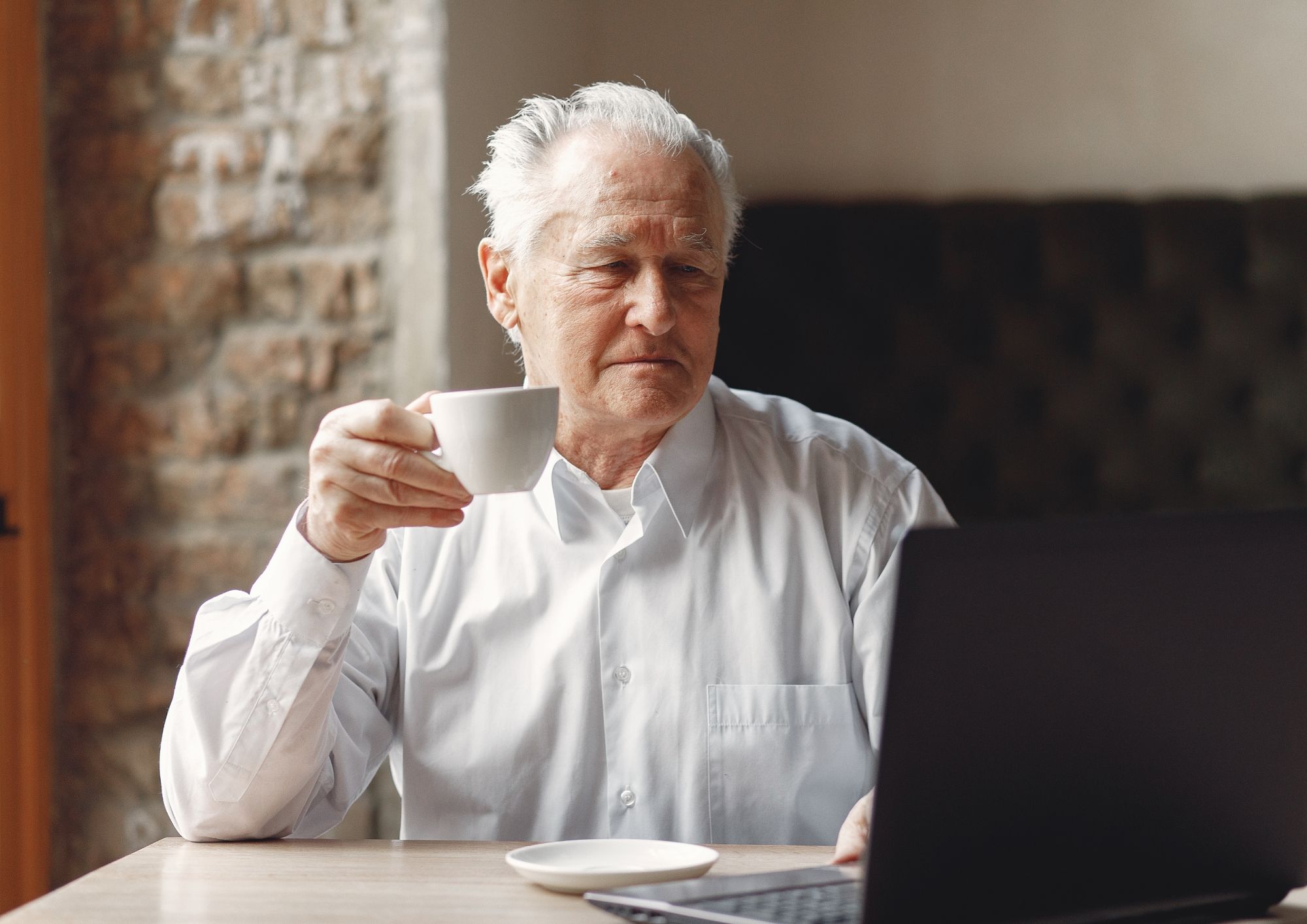 Äldre man i vit skjorta sitter vid ett bord med en kopp kaffe och tittar på en laptop.