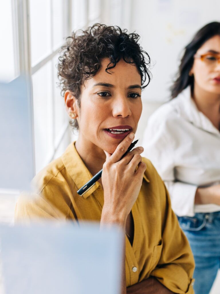 Two women in a modern office brainstorming ideas using sticky notes on a glass wall.