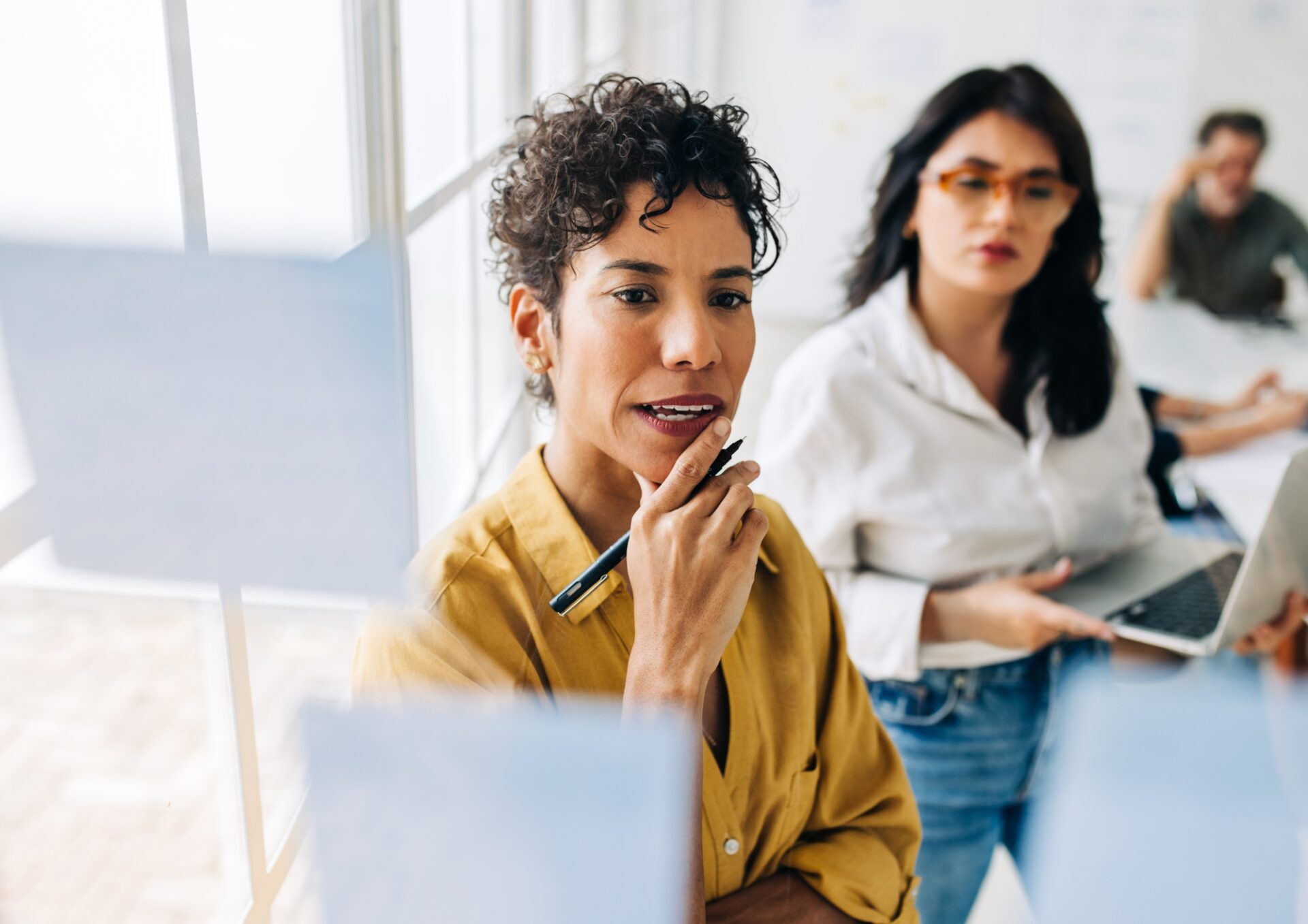 Two women in a modern office brainstorming ideas using sticky notes on a glass wall.