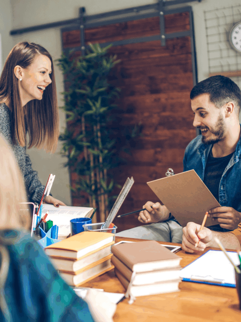 Fyra unga vuxna sitter runt ett bord och samarbetar i en avslappnad kontorsmiljö. De skrattar och diskuterar anteckningar med böcker och papper framför sig.