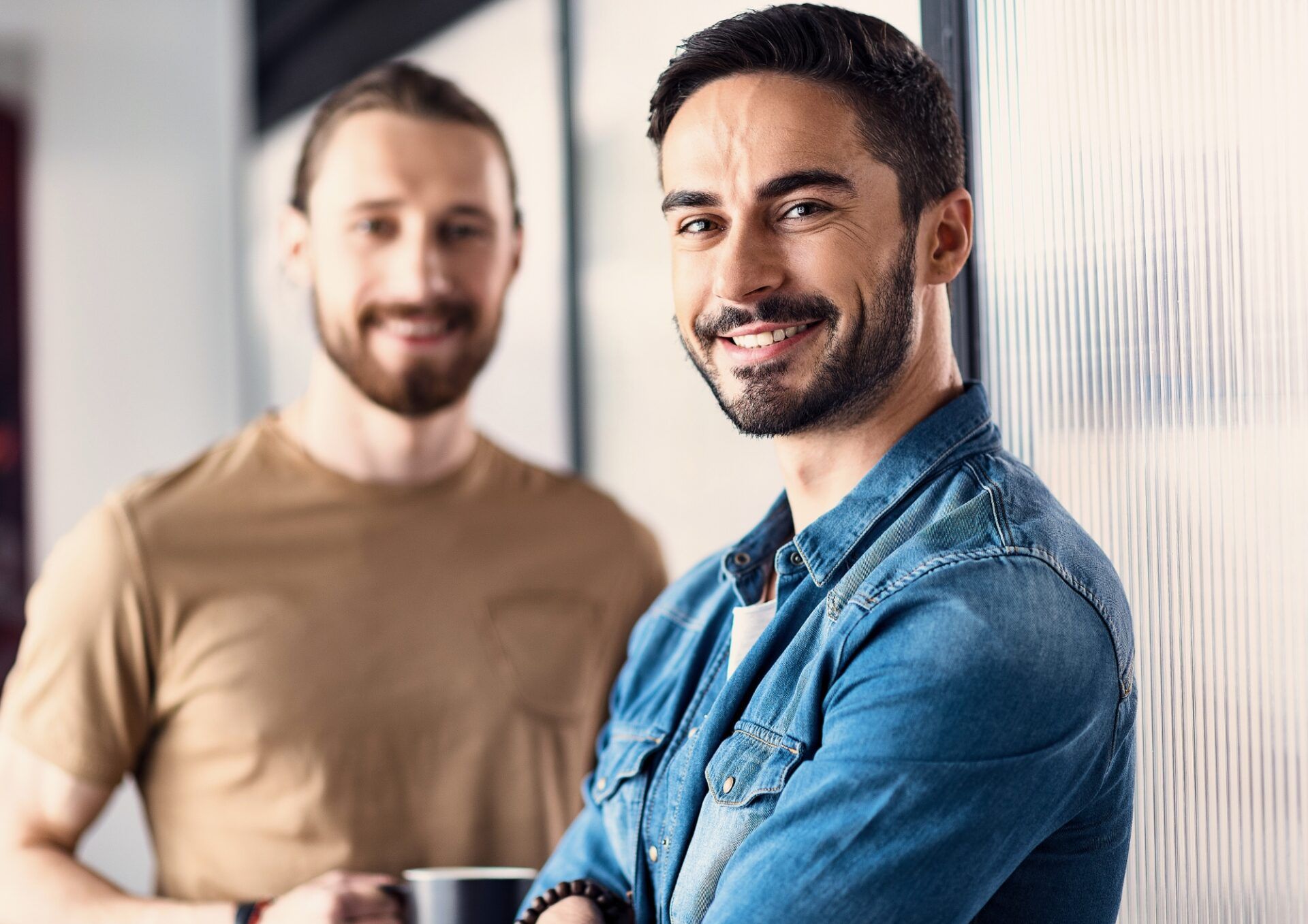 Person som tittar in i kameran iklädd blå denimskjorta i förgrunden. I bakgrunden en person som håller i en mugg och är iklädd en beige t-shirt