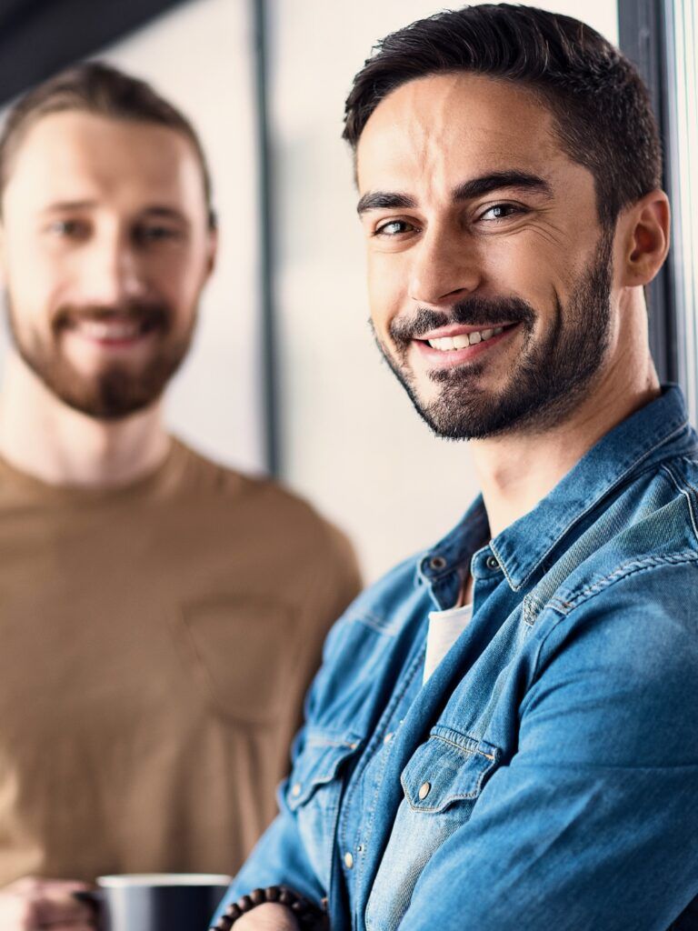 Person som tittar in i kameran iklädd blå denimskjorta i förgrunden. I bakgrunden en person som håller i en mugg och är iklädd en beige t-shirt