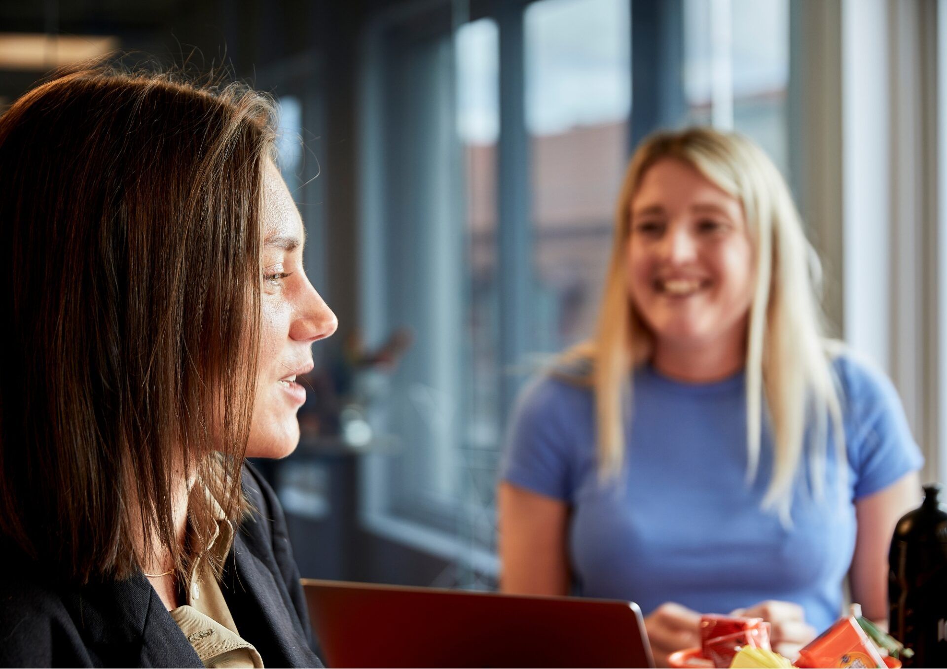 Martina Myrén syns i profil. Martina sitter och pratar och i bakgrunden sitter Ellinor Hellberg och ler stort mot Martina. De befinner sig i en kontorsmiljö.