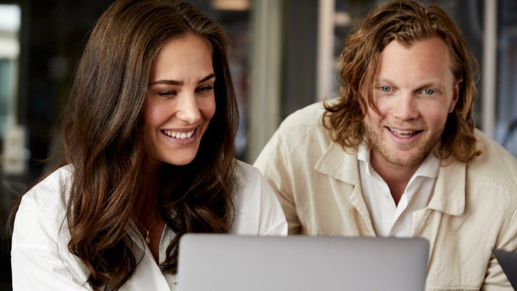 Kajsa Bengtsson och Charles Sinclair med leende ansikten tittar på en laptopskärm, i en ljus kontorsmiljö.