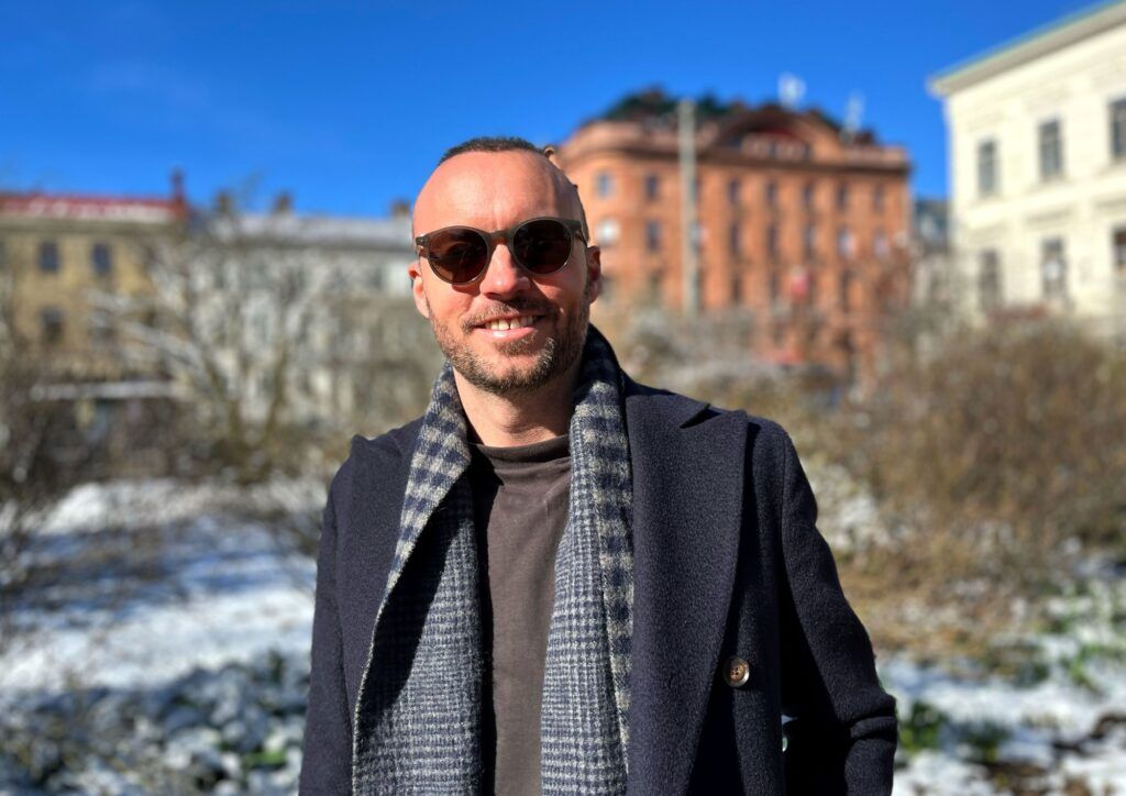 Foto på Niklas Kekonius utomhus. Han bär solgrasögon och i bakgrunden syns stenhus och blå himmel.